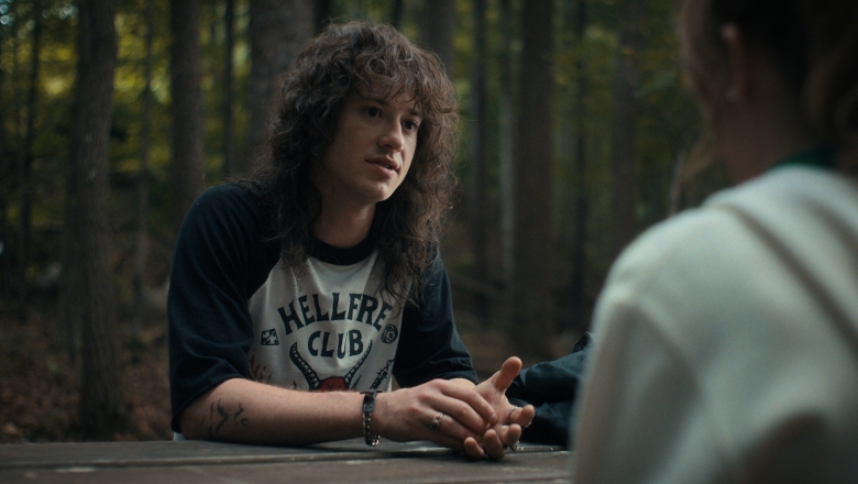 A young man with long, messy brown hair sitting at a picnic table in the woods; still from Stranger Things 4.
