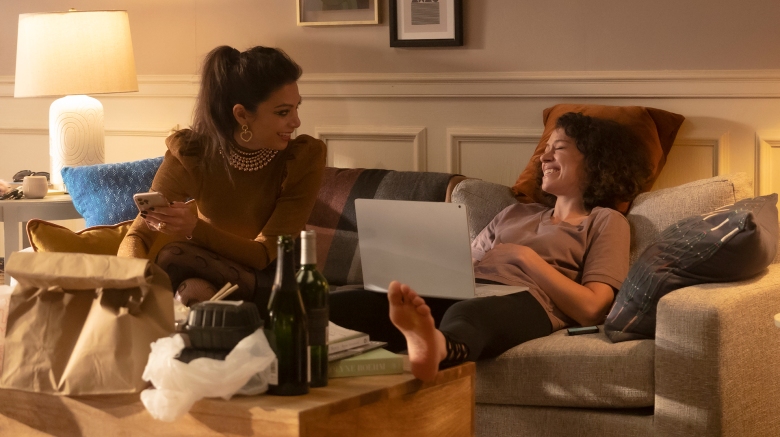 Two women sit on a couch, talking and laughing, with food and drinks on the coffee table in front of them; still from She-Hulk: Attorney at Law