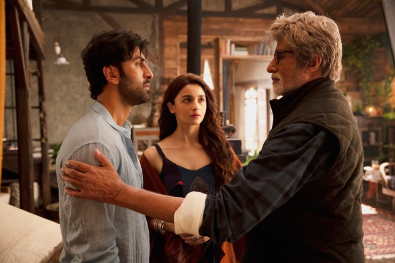 A gray-haired older man in glasses, grabs a younger man in a grey long-sleeve by the arms, while a woman with long brown wavy hair wearing a black dress watches between them; still from Brahmastra.