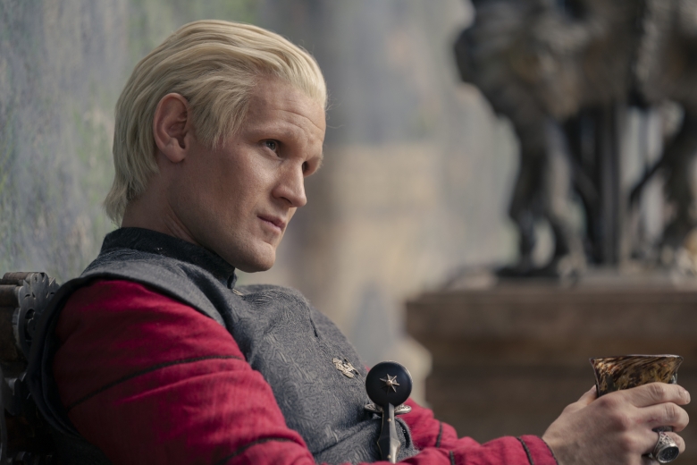 A man in a red tunic and gray vest with slicked back blonde hair, in a medieval setting; still from House of the Dragon