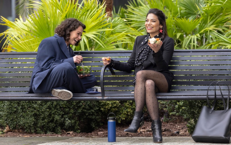 Two women sitting on a bench in front of bright green foliage, one of them sitting cross-legged in an overlarge suit and the other with her legs crossed, in a leather jacket and boots; still from She-Hulk.
