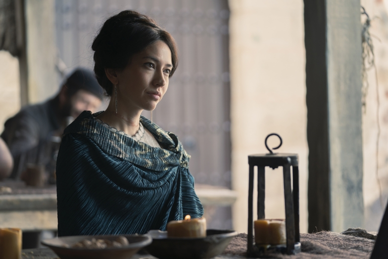 Medium shot of a seated woman with brown, baided hair and wearing a teal shawl; still from House of the Dragon