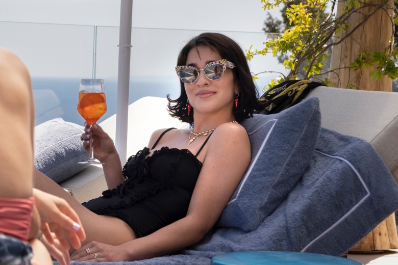 A woman with short, dark hair wearing a black one-piece swimsuit and sunglasses while laying in a pool chair and sipping an aperol spritz by the ocean; still from The White Lotus Season 2.