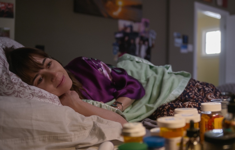 A sick woman laying in bed wearing a purple robe, her bedside table covered in prescription bottles; still from Dead to Me