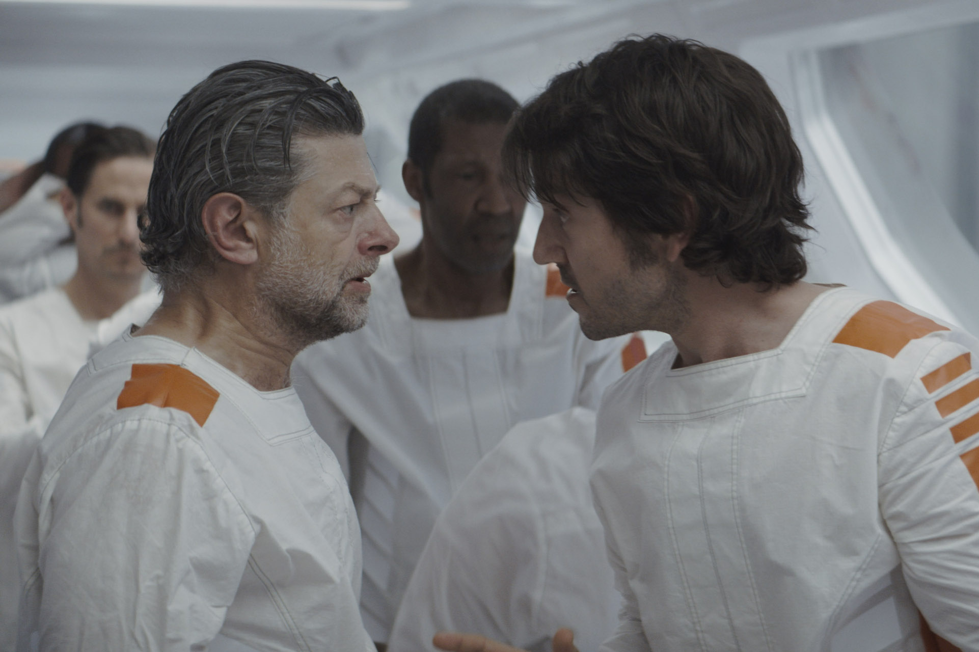 Two men in white prison garb face each other, next to a line of other men; still from Andor.