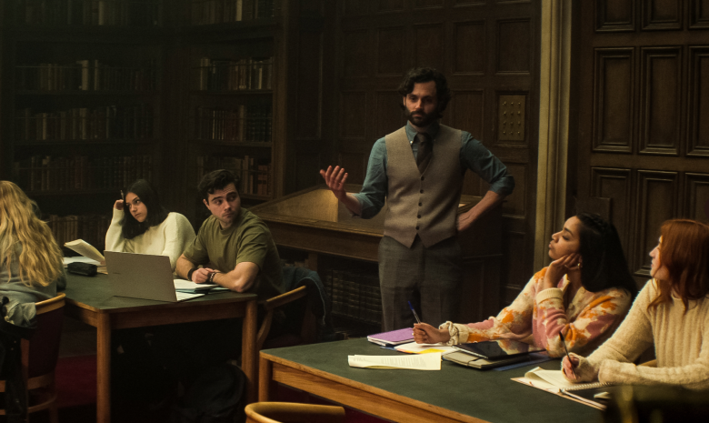 A man in a blue shirt and gray vest lectures a small class of college students in a library; still from You Season 4.
