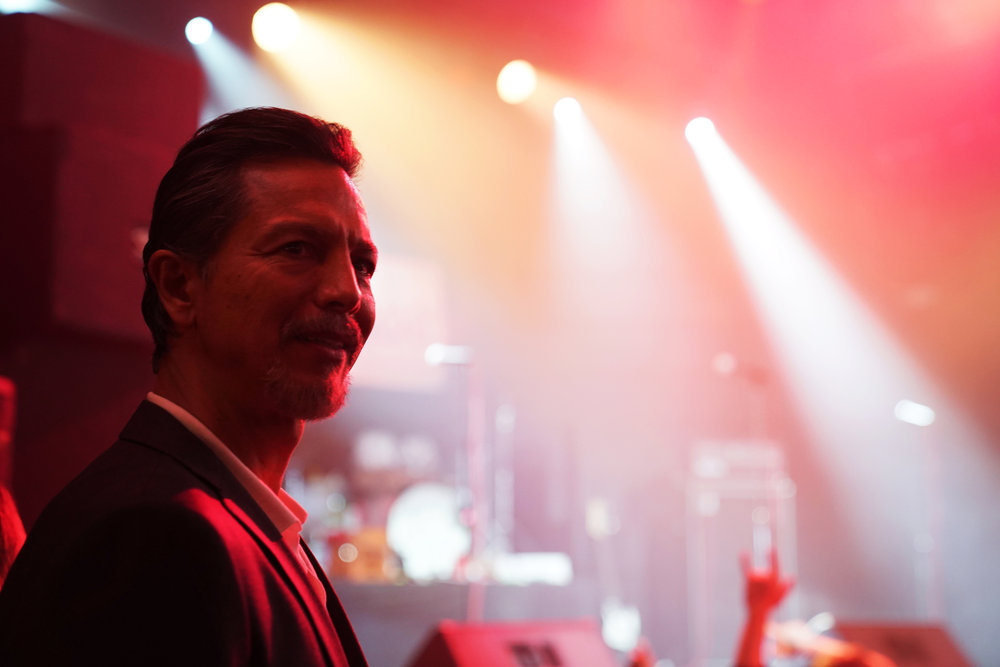 A suited man with a goatee stands in the foreground with a lit, empty stage and concert crowd in the background; still of actor Benjamin Bratt in Poker Face