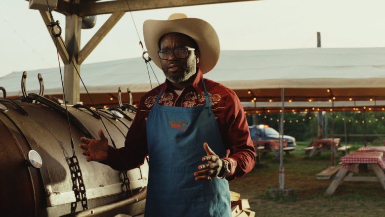 A bespectacled man in a cowboy hat and apron uses his hands to explain something; still of actor Lil Rel Howery from Poker Face