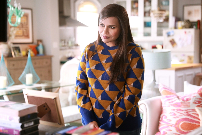 A woman with long hair wearing a brown and navy blue patterned sweater, standing in a brightly-lit and colorful office; still from The Mindy Project